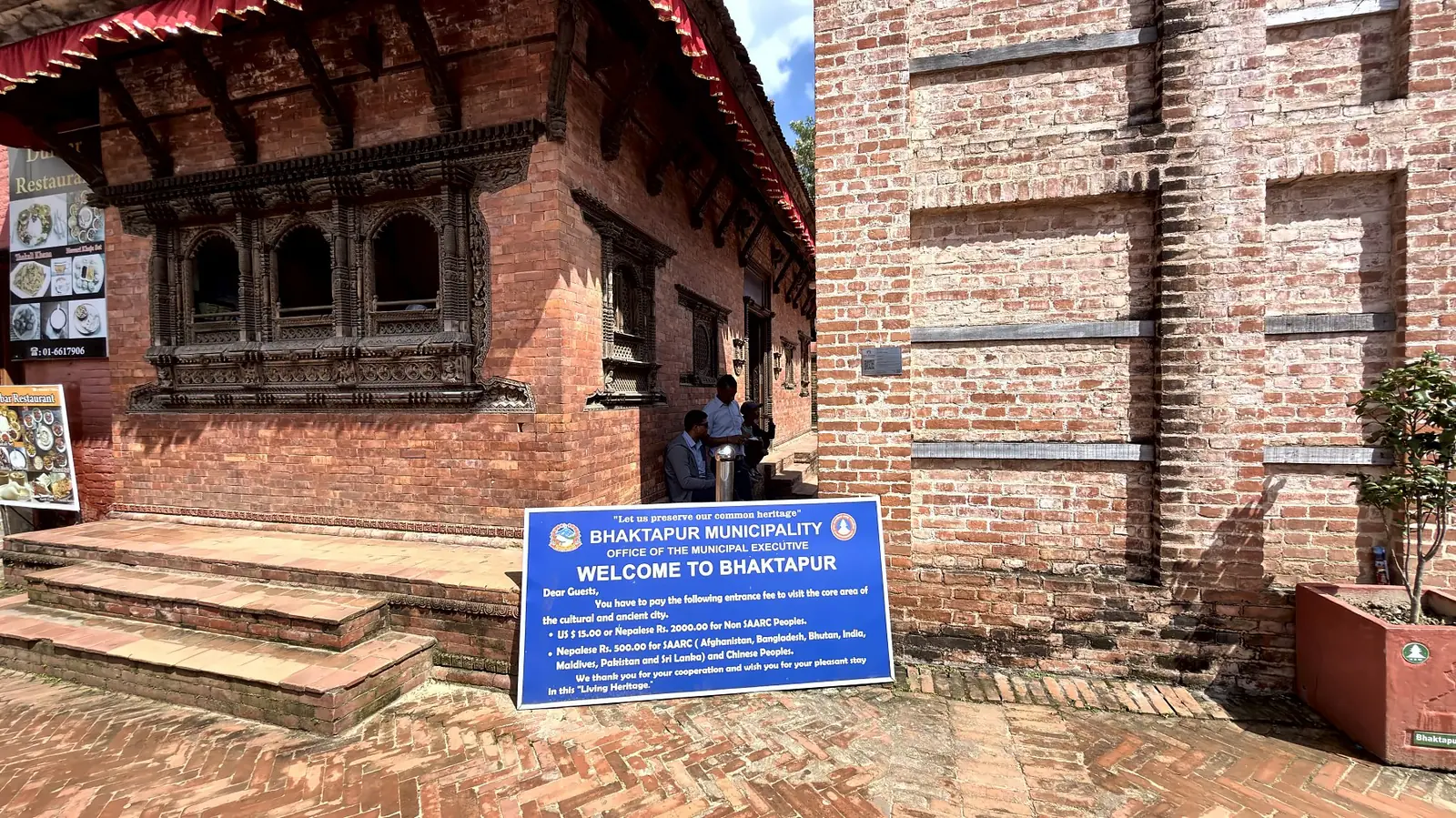 Blue Bhaktapur Municipality welcome board for visitors outside a carved brick building